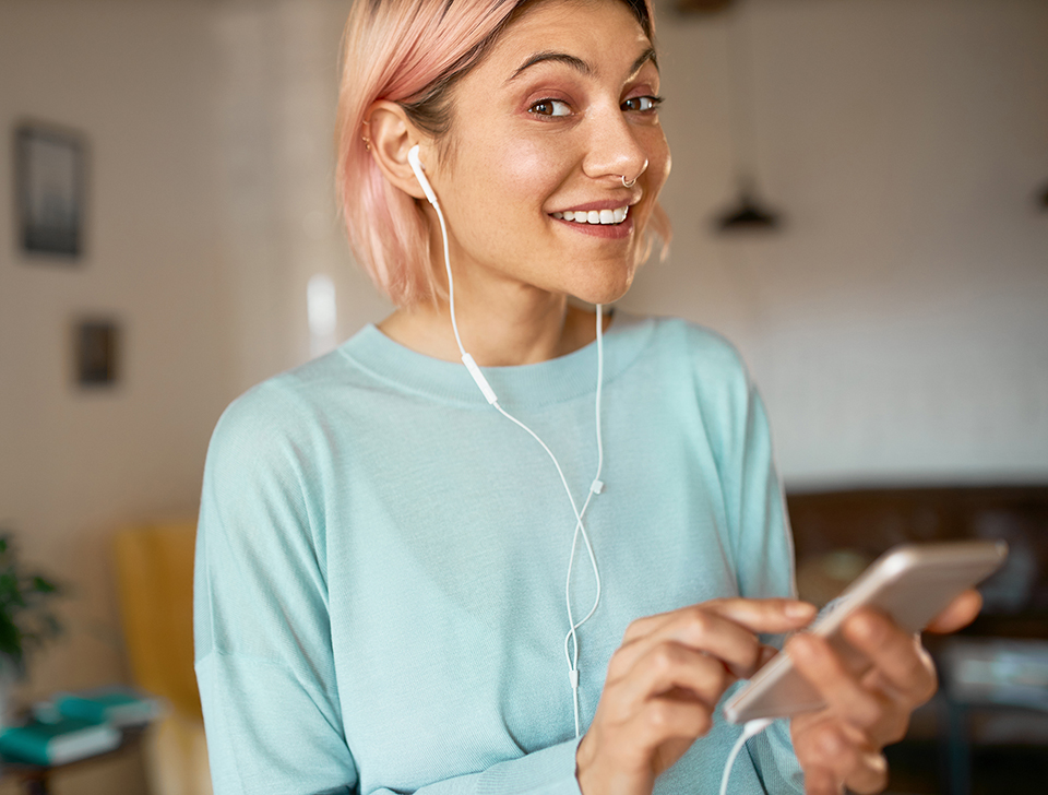 Positive cute student girl having fun indoors after college using headset while chatting online via video chat on her smart phone, enjoying music, browsing newsfeed on social networks, smiling